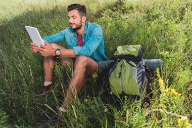 Dijital tablet kullanma ve sırt çantası ile çimenlerin üzerinde oturan turist