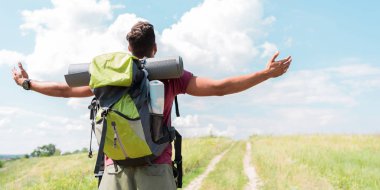 back view of traveler with backpack standing with outstretched hands on green meadow with cloudy sky