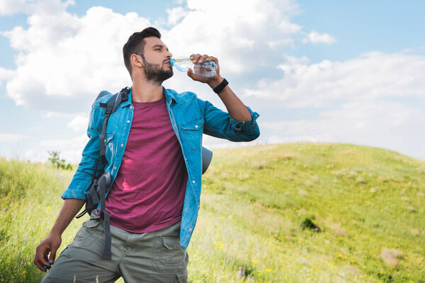 handsome traveler with backpack drinking water on summer meadow