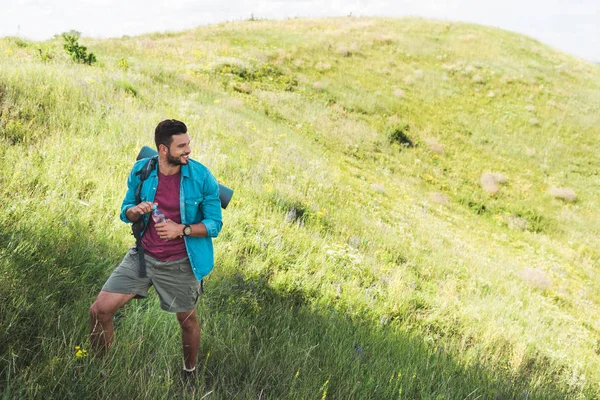 traveler with backpack holding bottle of water and standing on summer meadow