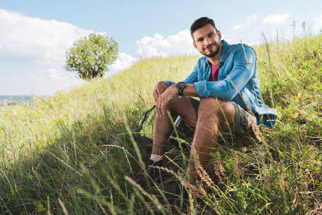 handsome smiling traveler sitting on green summer meadow
