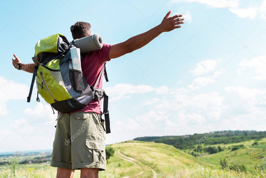 tourist with backpack standing with outstretched hands on summer meadow with cloudy sky