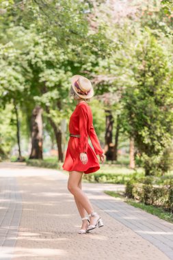 stylish girl in red dress and straw hat standing on road in park clipart