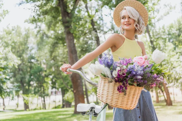 attractive girl holding bike with basket of flowers in park