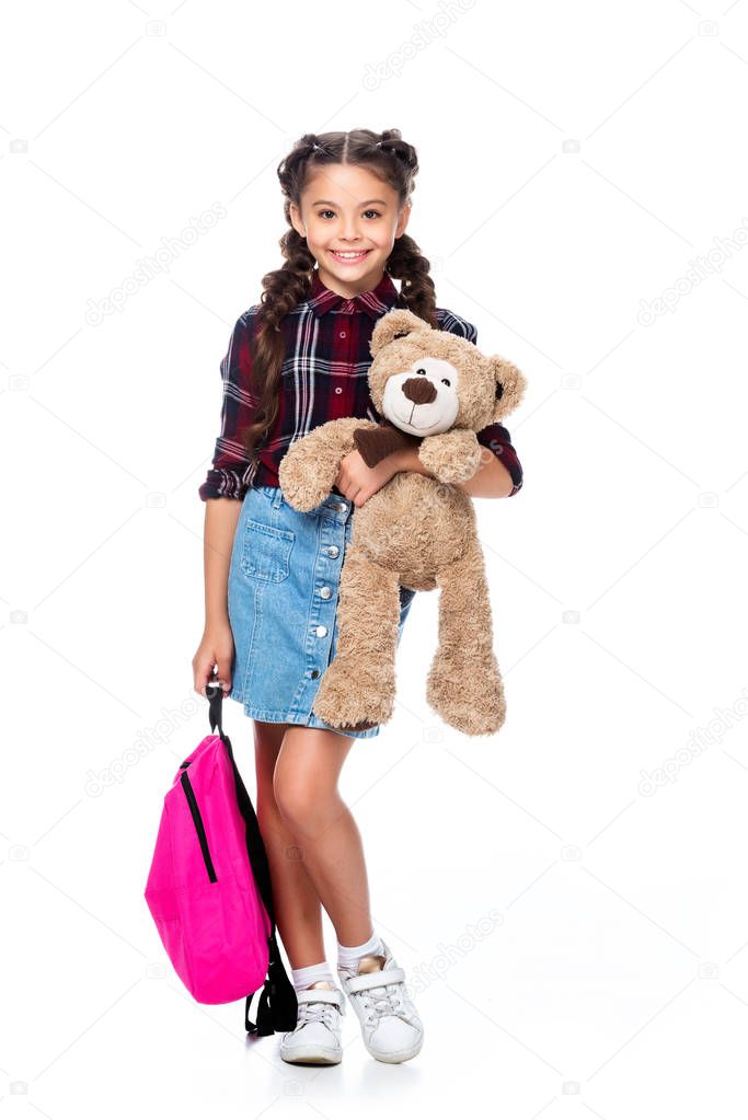 smiling schoolchild holding teddy bear and backpack isolated on white