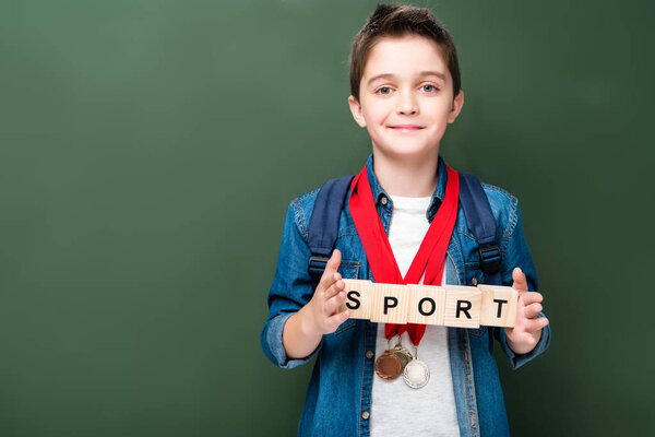 schoolboy with medals holding wooden cubes with word sport near blackboard