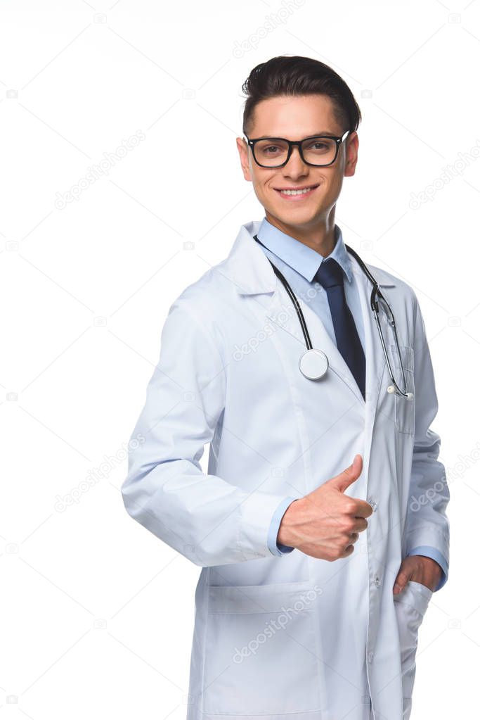 smiling young doctor showing thumb up and looking at camera isolated on white