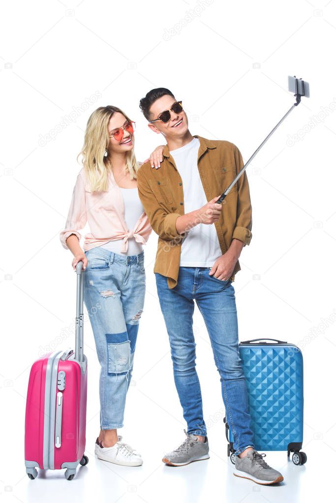 young couple with suitcases taking selfie with smartphone from monopod isolated on white