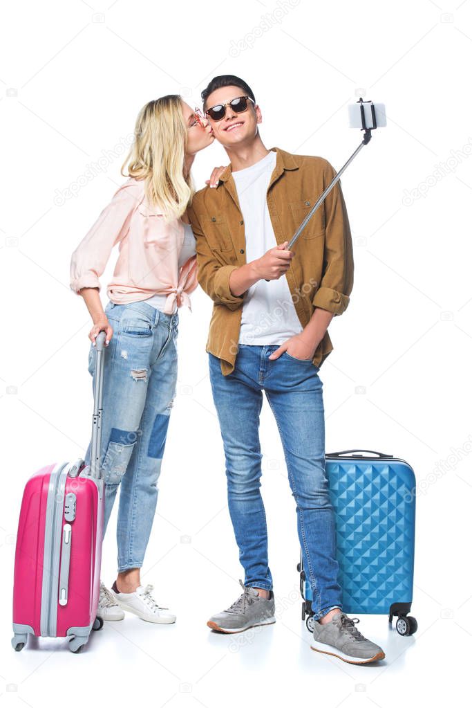 young couple with suitcases taking selfie from monopod while woman kissing her boyfriend isolated on white