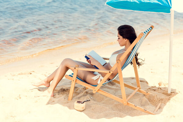 beautiful girl dearing book on beach chair with coconut cocktail on sand