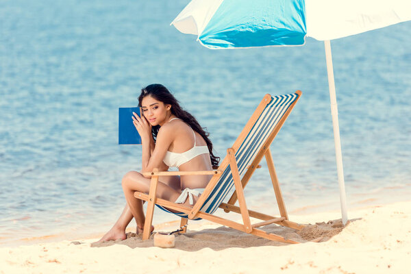 attractive girl in white bikini with book on beach chair near sea