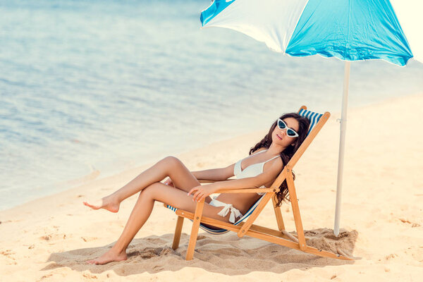 woman in sunglasses and bikini relaxing on beach chair under umbrella near the sea