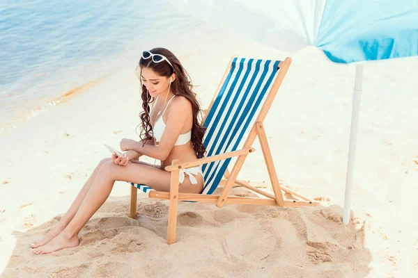 魅力的な女性のスマート フォンを使用して ビーチ リゾートで海の近くの太陽の傘の下で椅子が休んで  — 無料ストックフォト