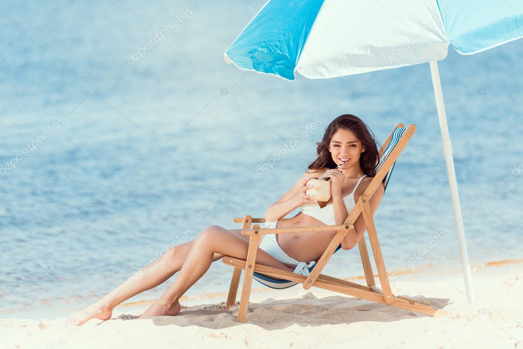 happy young woman drinking cocktail in deck chair under sun umbrella near sea