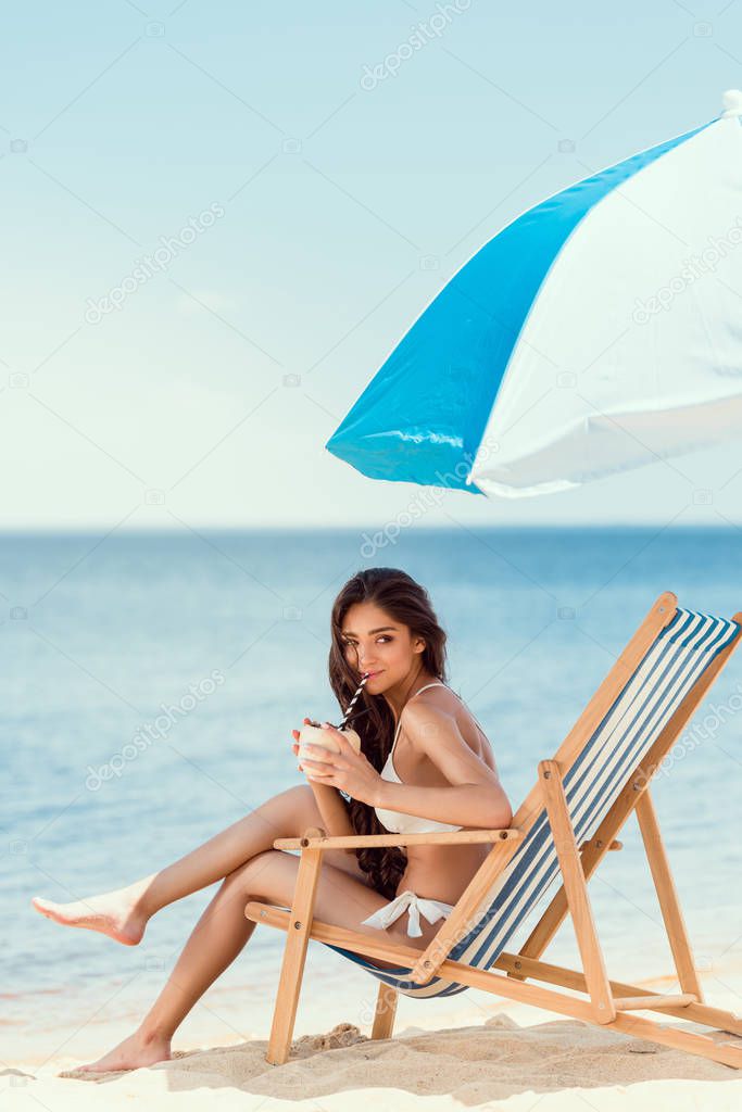 young woman drinking cocktail in beach chaise longue under sun umbrella