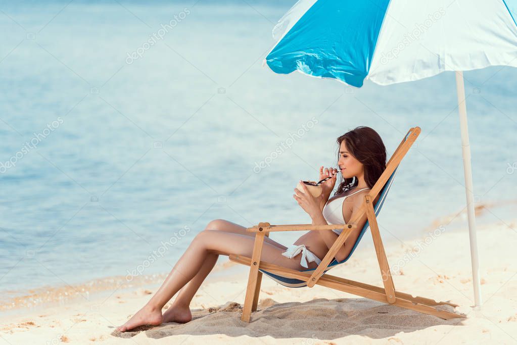 attractive girl in bikini drinking coconut cocktail in beach chair under umbrella