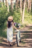 zadní pohled na dívku v slaměný klobouk a bílých šatech chodit s kolo a květiny v proutěném koši