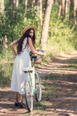 hasır şapka ve beyaz elbise ile bisiklet ormandaki izinde yürüyen genç kadın