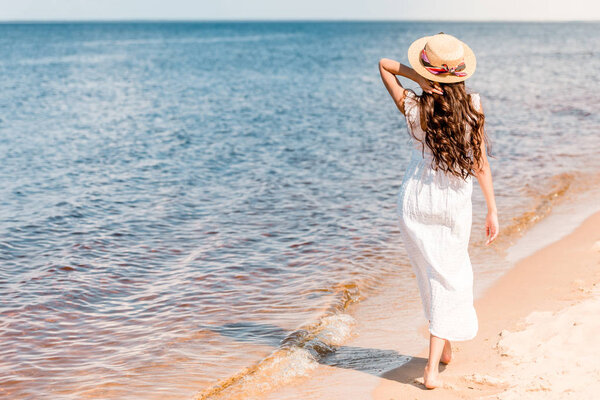 back view of woman in straw hat and white dress walking on beach near sea