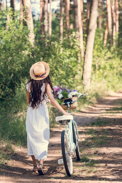 back view of girl in straw hat and white dress walking with bicycle and flowers in wicker basket