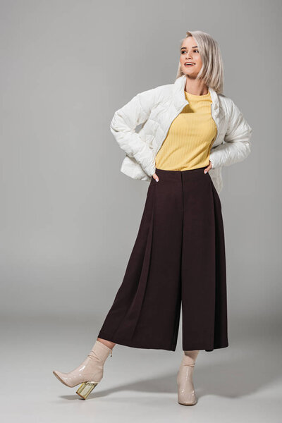 счастливая модная девушка в осеннем наряде позирует с руками на талии на сером фоне
 