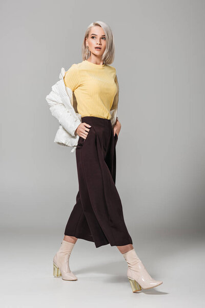 молодая модель в стильном наряде, позирующая руками в карманах на сером фоне
 