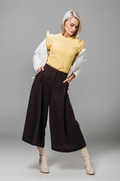 модная молодая женщина в стильном наряде позирует с руками в карманах на сером фоне
 