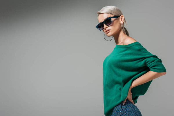 fashionable young woman in sunglasses posing with hands on waist isolated on grey background