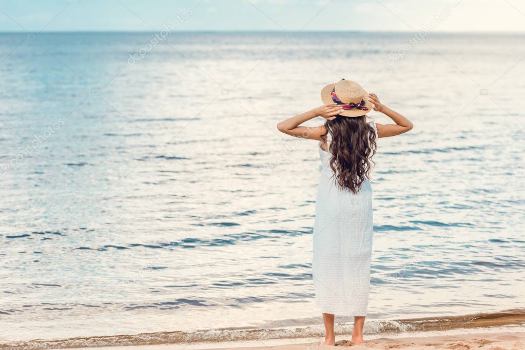 rear view of woman in straw hat and white dress standing on beach and looking at sea