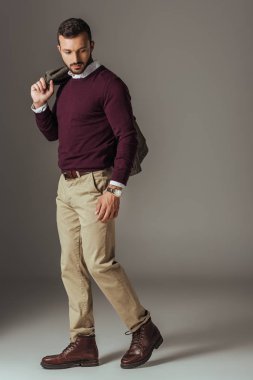 fashionable man posing in beige pants and burgundy sweater with autumn jacket on shoulder, on grey clipart