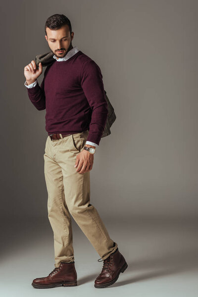 fashionable man posing in beige pants and burgundy sweater with autumn jacket on shoulder, on grey