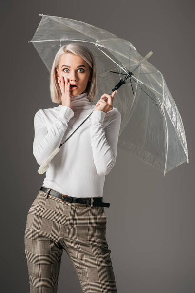 elegant shocked girl in white turtleneck posing with fashionable transparent umbrella, isolated on grey