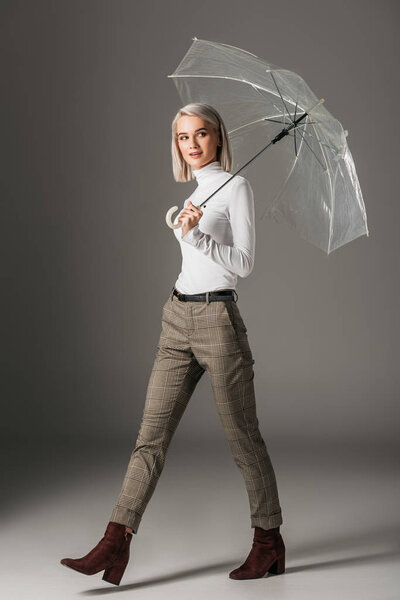 elegant blonde girl in white turtleneck and grey pants walking with transparent umbrella, on grey