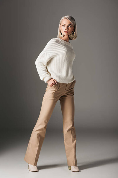 elegant blonde woman posing in white sweater and beige pants, on grey
