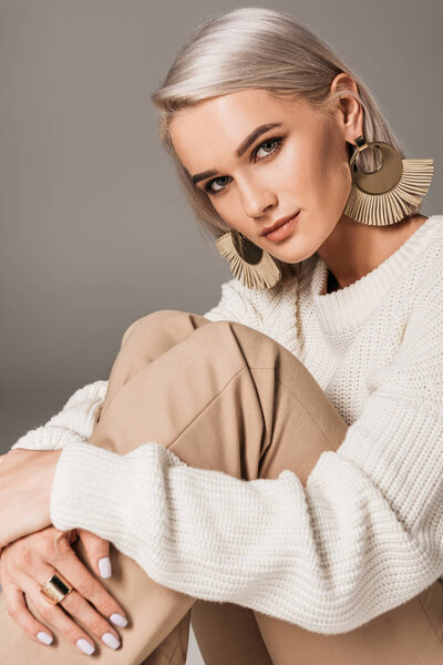 elegant stylish woman in white sweater and big earrings looking at camera, isolated on grey