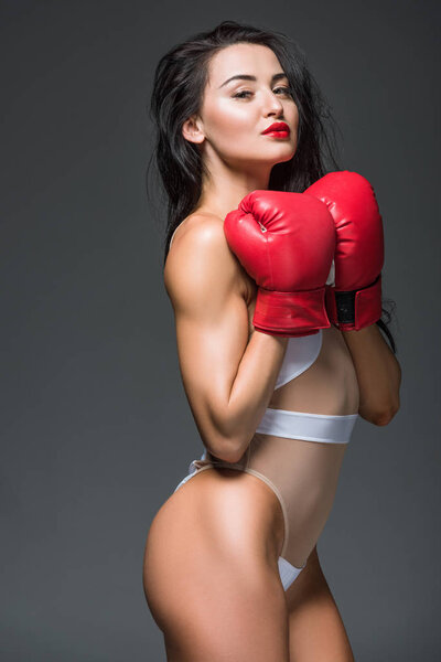 side view of sexy sportive woman in white swimwear and boxing gloves isolated on grey