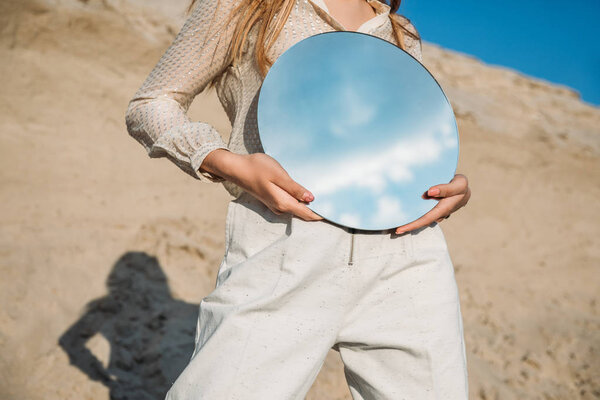 cropped view of stylish girl holding mirror with reflection of blue sky with clouds