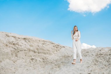 beautiful woman in white fashionable clothes posing on sand dune with blue sky clipart