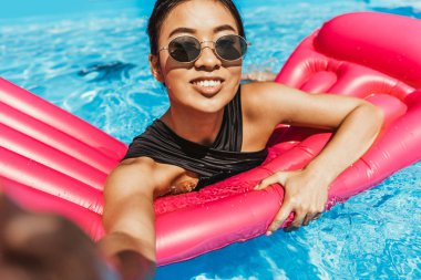 smiling asian girl in sunglasses taking selfie on inflatable mattress in swimming pool clipart