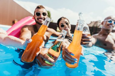 multiethnic friends clinking with bottles on beer in swimming pool