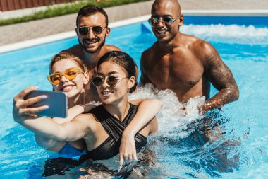 güneş gözlüğü Smartphone cep telefonu ile yüzme havuzunda selfie alarak çok ırklı arkadaşlar