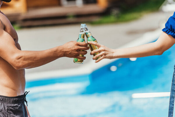 cropped view of couple clinking with bottles of beer at poolside
