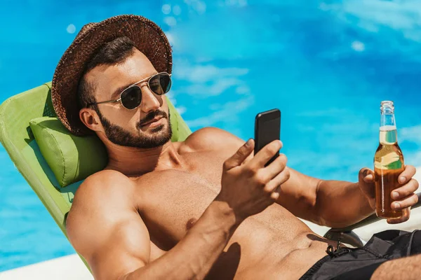 男子在太阳镜使用智能手机和控股瓶啤酒日光浴在池畔 — 免费的图库照片