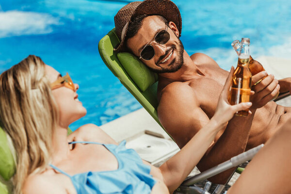 smiling couple clinking with bottles of beer while lying on sunbeds near swimming pool
