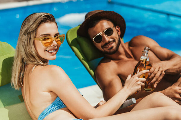 smiling couple in sunglasses clinking with bottles of beer while lying on sunbeds
