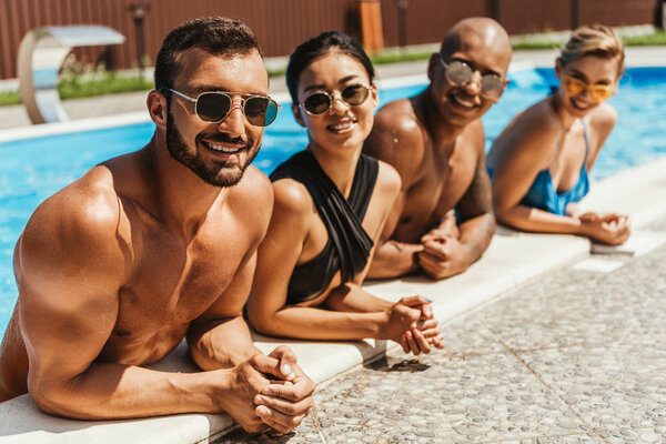 multiethnic friends in swimsuits and sunglasses resting in swimming pool