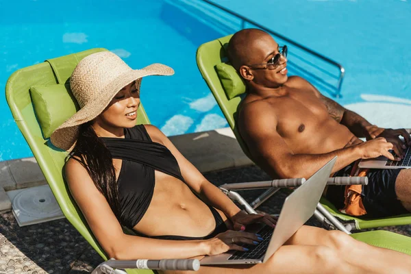 多文化共生のカップル プールサイドのサンベッドでリラックスしながらノート パソコンを使用して — ストック写真