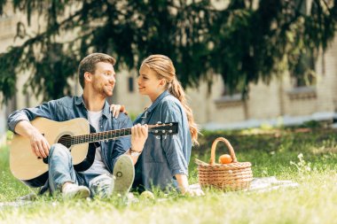 çekici bir genç adam gitar gülümseyen kız arkadaşı için piknik sırasında parkta oynarken