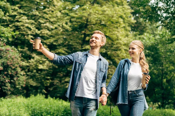 快乐的年轻夫妇与纸杯咖啡走在一起公园 并指向某处 — 图库照片