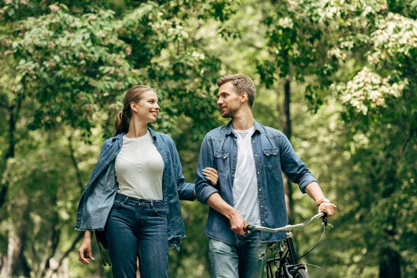 happy young couple in denim shirts with bicycle walking by park